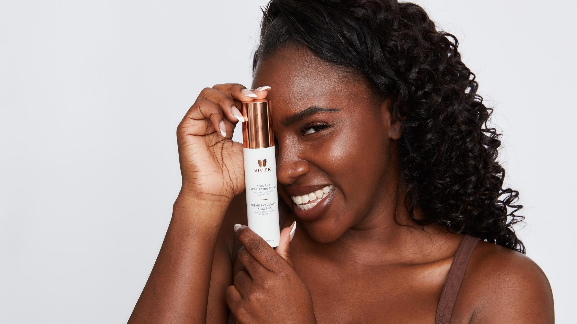 Person smiling while holding a tube of Vivier AHA BHA Exfoliating Cream against a soft neutral background.