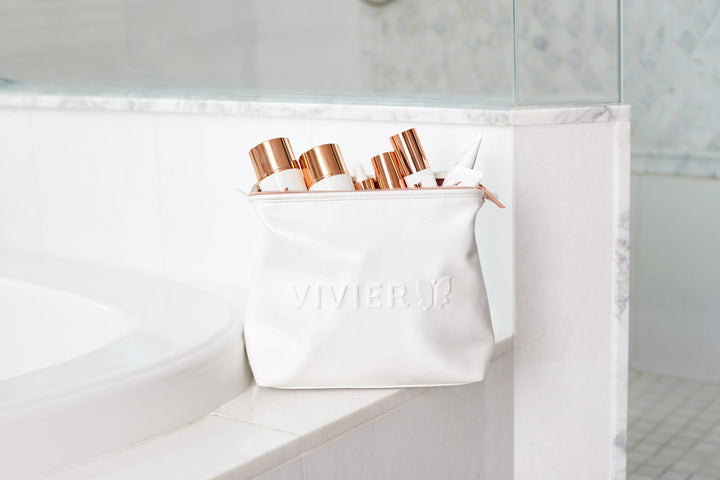 White Vivier skincare bag filled with products, placed on a bathroom counter beside a bathtub.