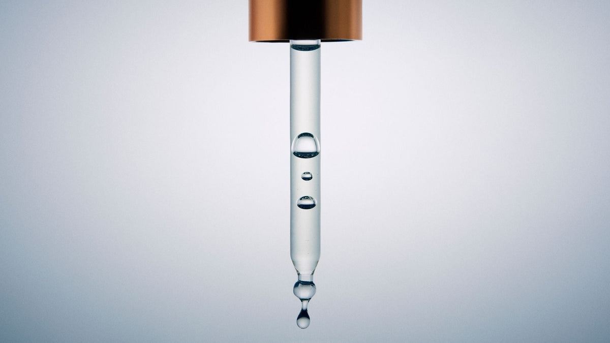 Close-up of a glass serum dropper containing clear skincare serum with suspended droplets against a soft grey background.